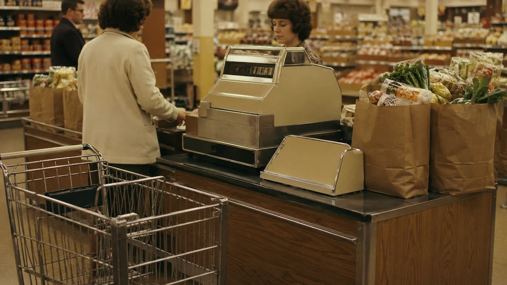 A 1970s supermarket checkout counter with paper grocery bags and a vintage cash register